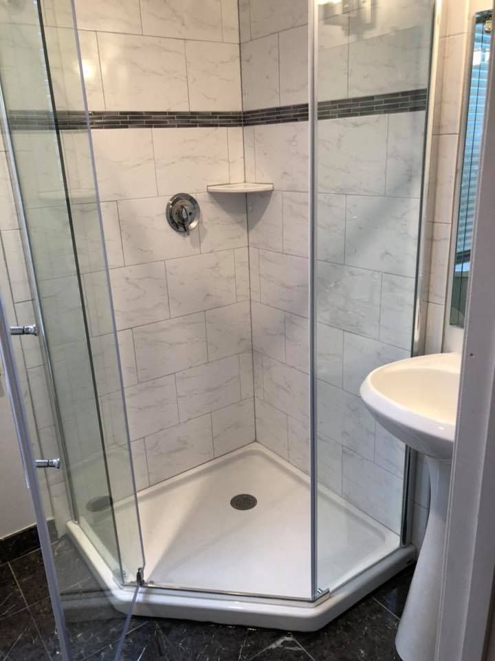 Corner shower with glass doors, white tile, small shelf, and sink on a pedestal.