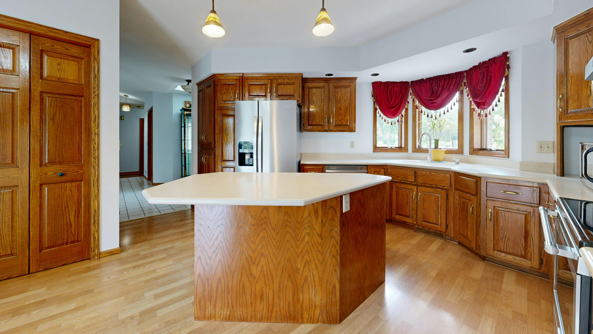 Kitchen with light wood cabinets, island, stainless steel refrigerator, and a bay window with red curtains.