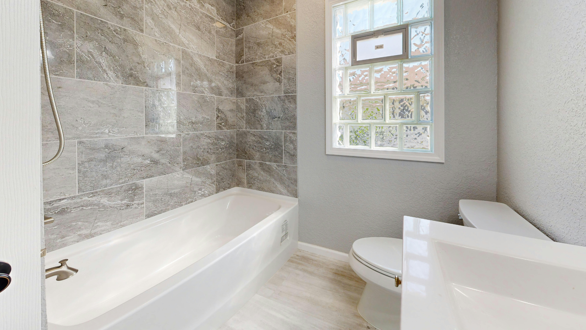 Bathroom with white tub, toilet, and sink. Grey tiled shower wall and window with glass blocks.