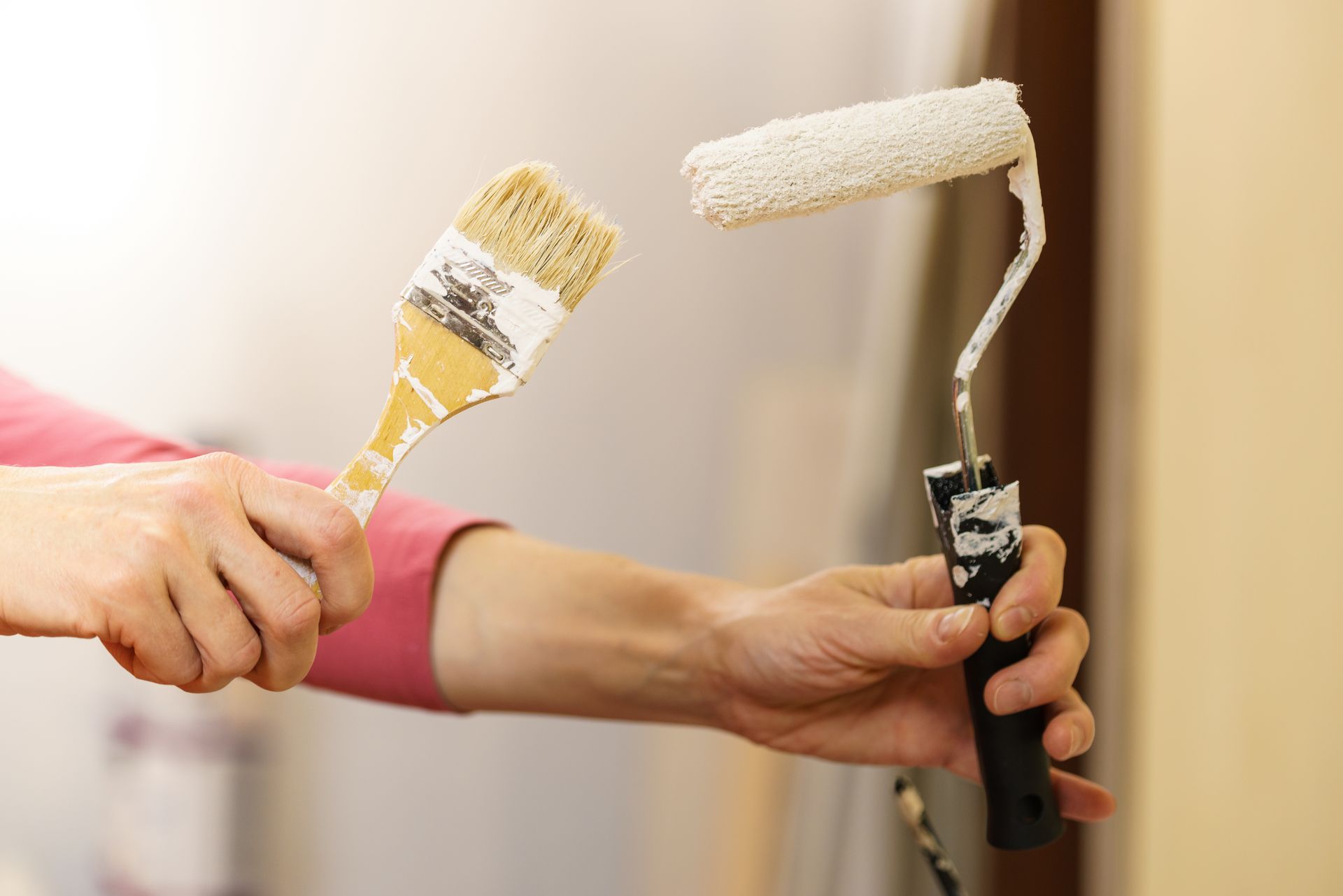 Hands holding a paint brush and paint roller, both with white paint, against a light-colored wall.