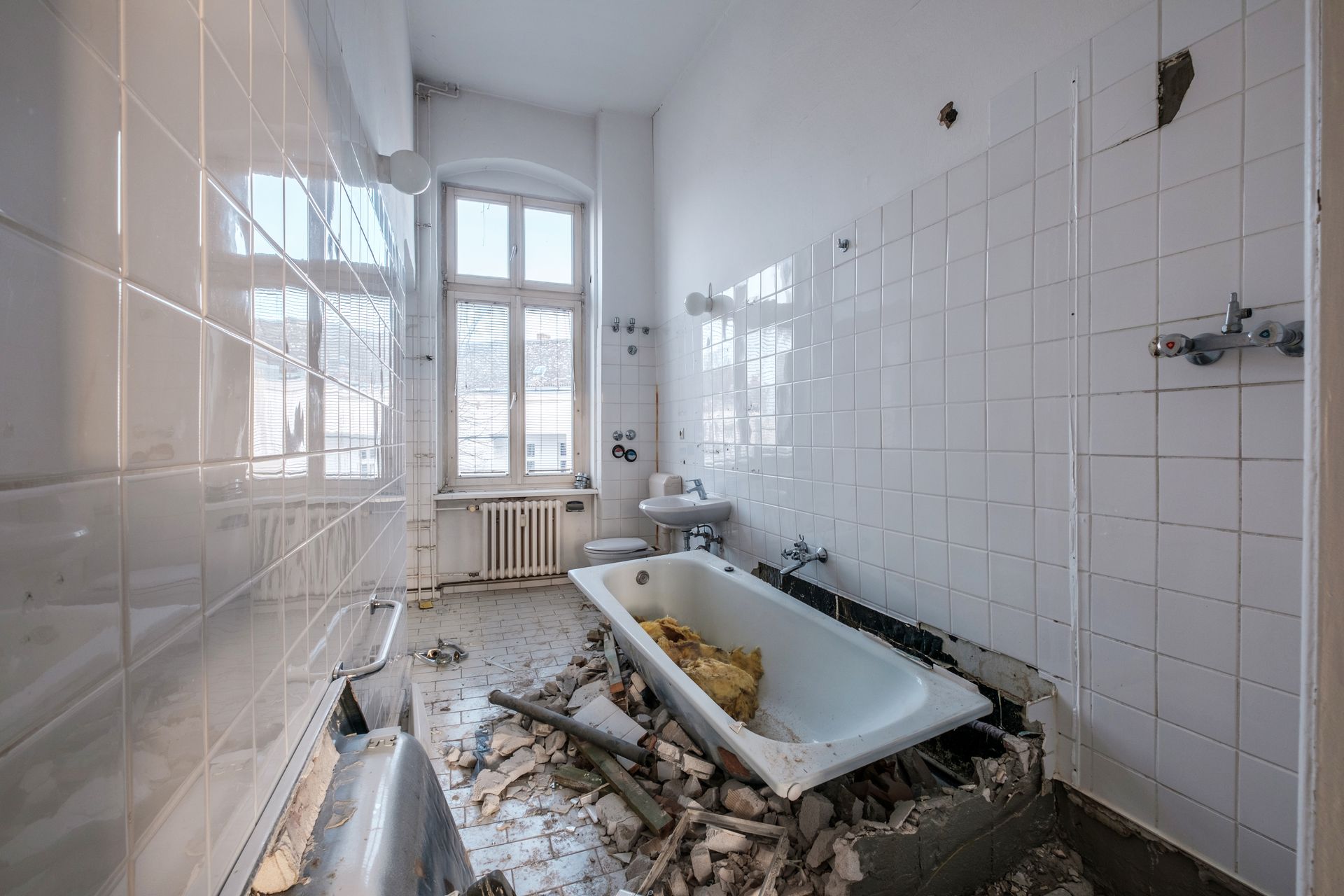 Bathroom in disrepair; white tile walls, a tub surrounded by debris, and a window.