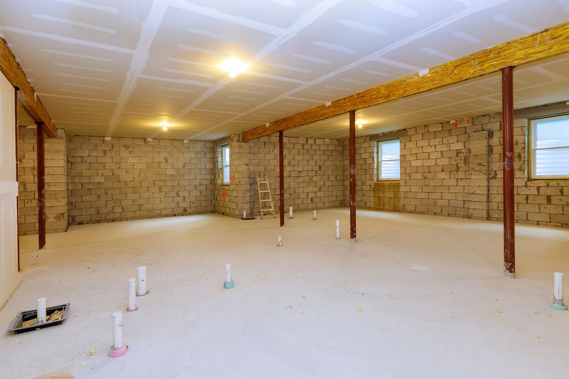 Unfinished basement with concrete block walls, support beams, drywall ceiling, and plumbing.
