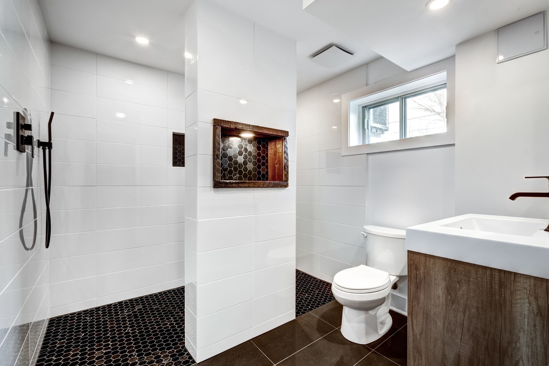 Modern bathroom with white tiled walls, black pebble shower floor, wooden vanity, and toilet.