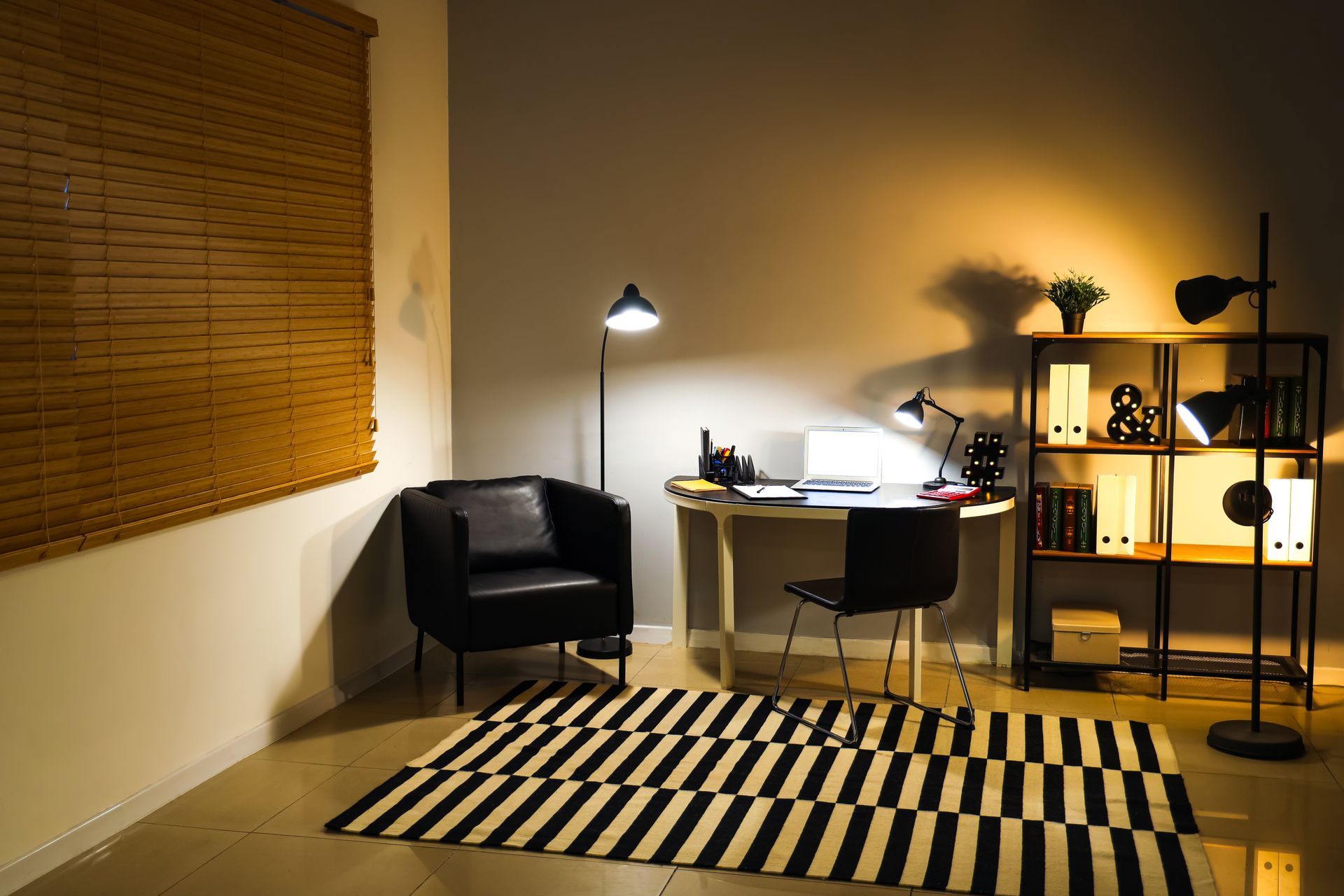 Cozy home office with desk, chair, black armchair, striped rug, bookshelf, and warm lighting.