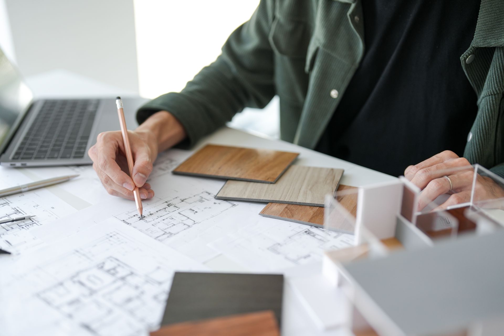 Person drawing on blueprint with pencil, wood samples and model home on desk. Laptop in background.