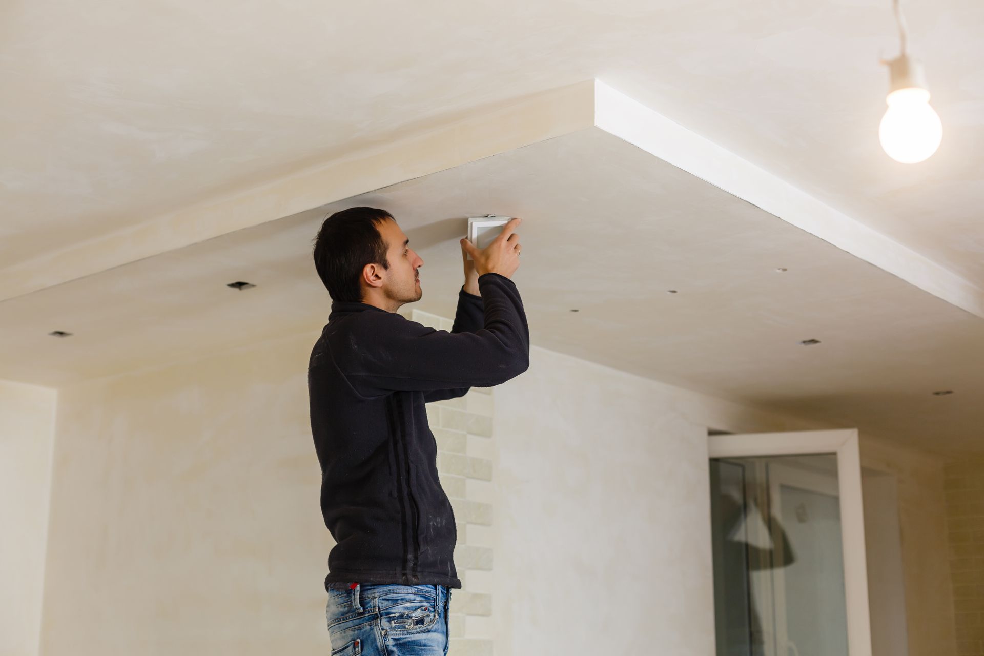 Man on stepladder installing a device on a white ceiling. Interior setting with a lightbulb.
