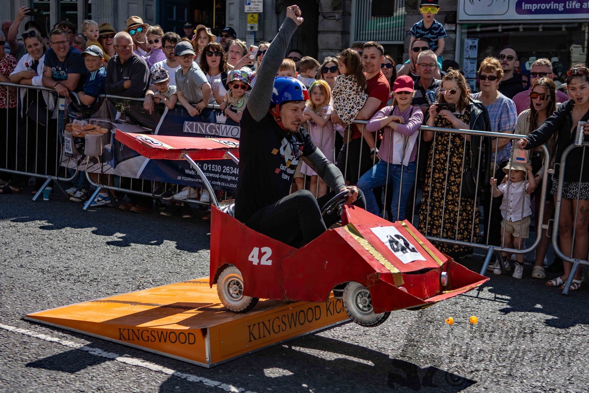 A man is driving a cardboard go kart on a street in front of a crowd.