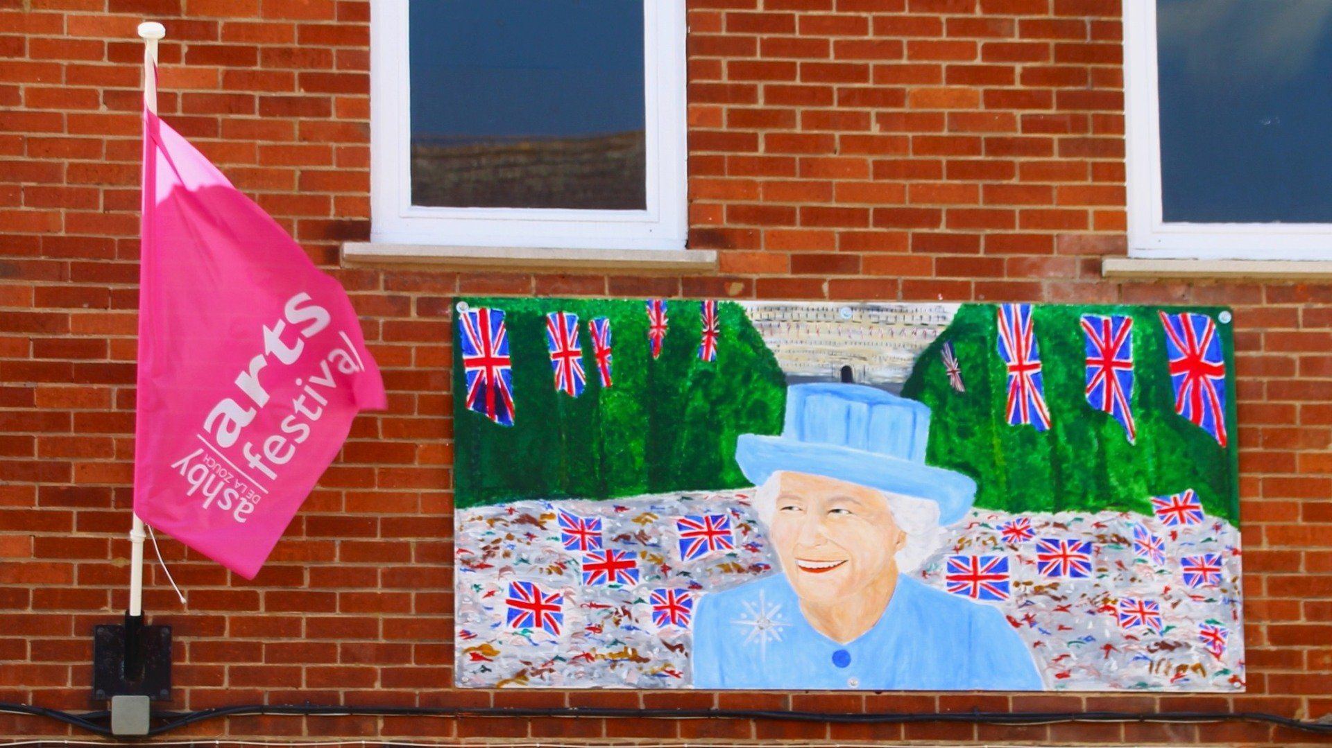 A brick wall with a painting of queen elizabeth and a pink arts festival flag
