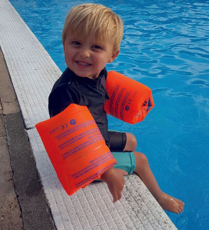 A young boy is sitting on the edge of a pool wearing orange arm floats