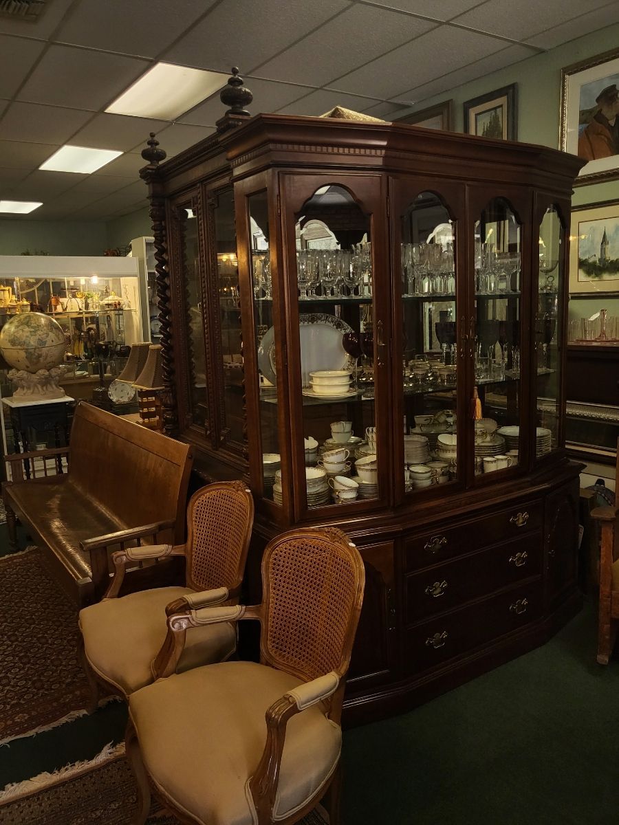 Wooden china cabinet with glass doors, drawers, and a decorative top, in a room with two chairs.