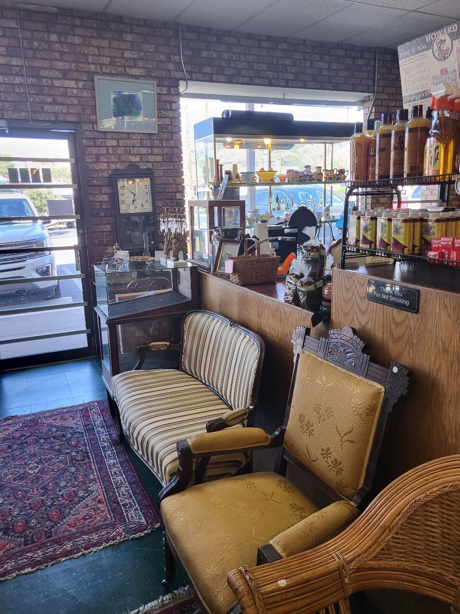 Cozy antique shop interior: brick wall, vintage furniture, rug, clock, display cases, and jars of goods near a window.