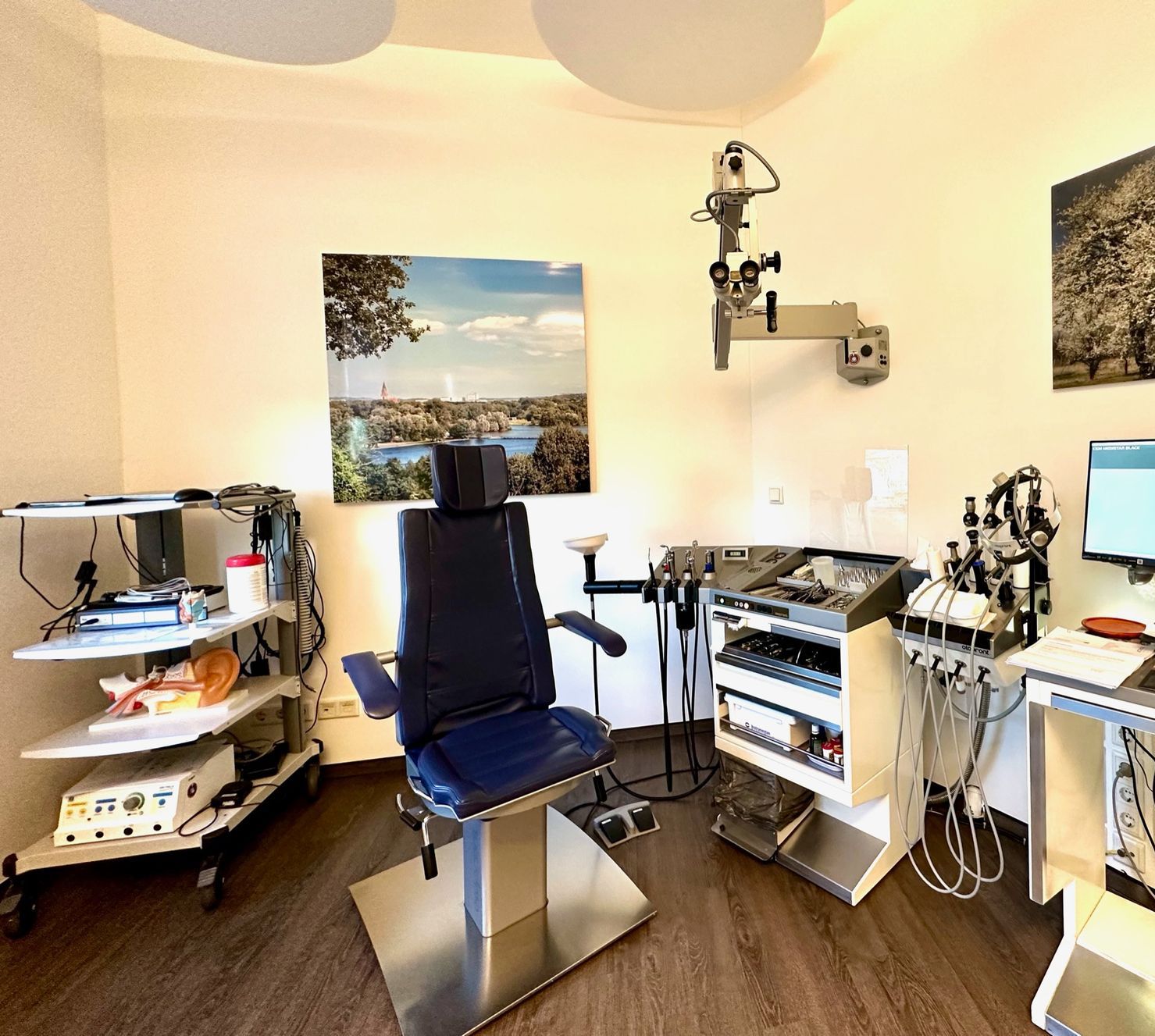 Das Bild zeigt das Behandlungszimmer der Praxis. An der Wand sind große Fotos von Neubrandenburg aufgegangen. In der Mitte steht ein blauer Behandlungsstuhl. Man sieht außerdem einige medizinische Geräte.