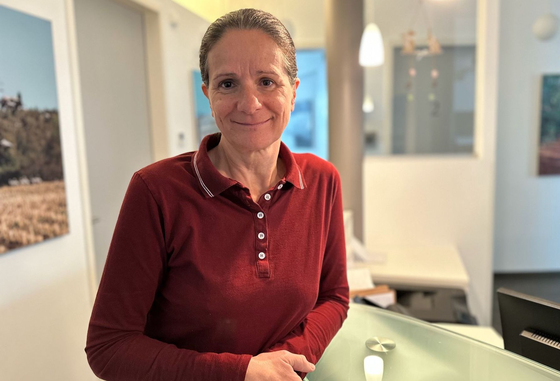 Das Bild zeigt die Doktorin Frau Silka Bohlmann in einem dunkelroten Oberteil. Sie schaut lächeln in die Kamera und lehnt am Tresen im Empfangsbereich .