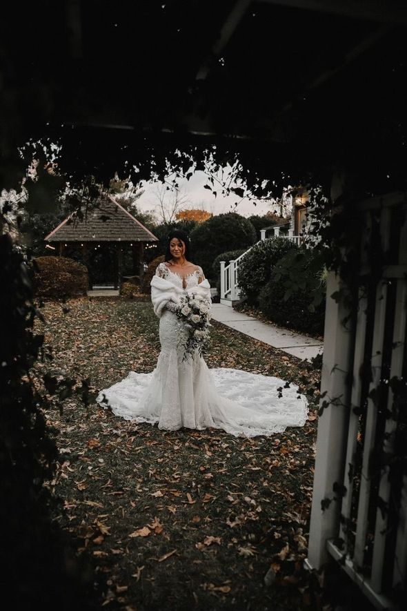 Bride in white dress gazes at groom in blue suit, holding bouquet; trees in background.