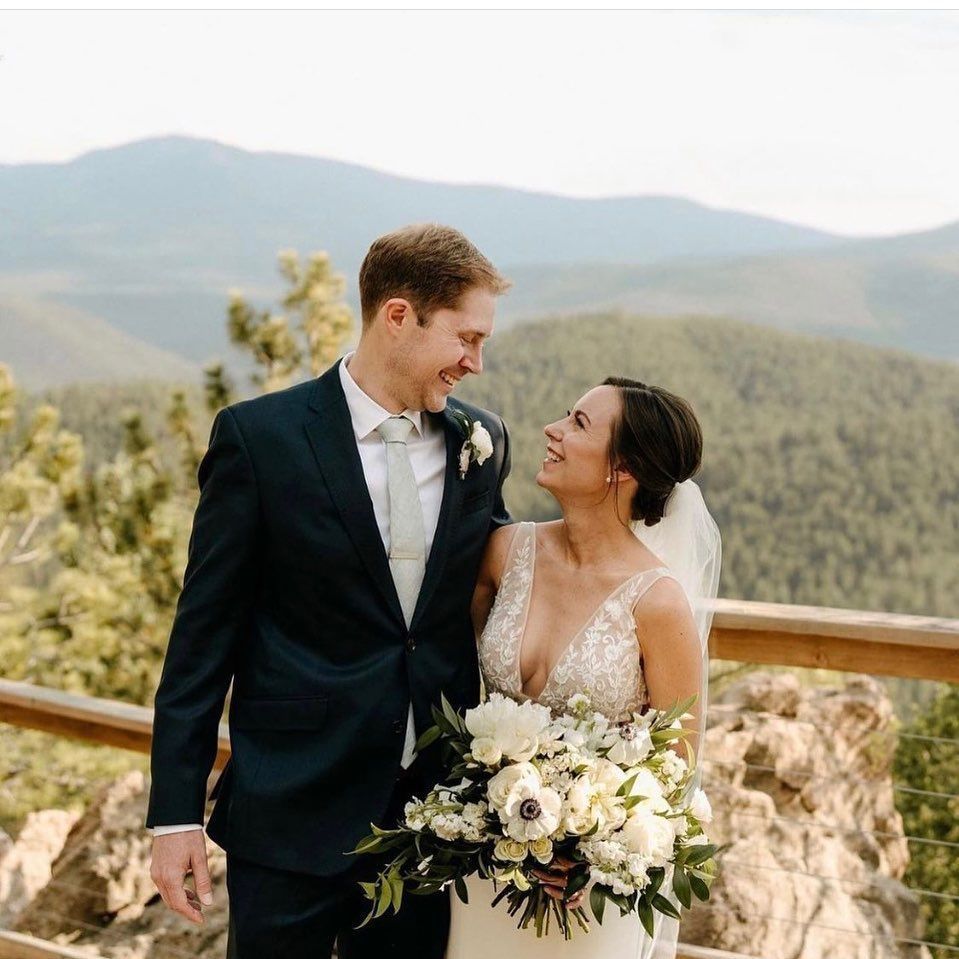 Bride and groom smiling at each other outdoors, bouquet in hand, against mountain background.
