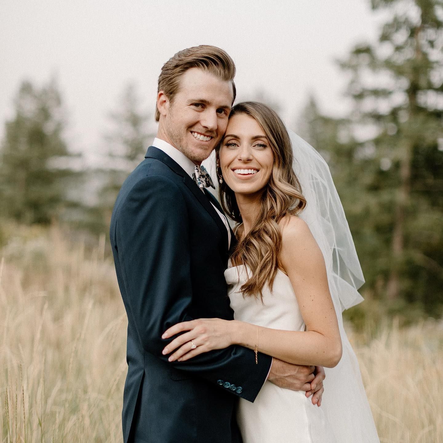 Wedding photo: Bride in white dress and groom in blue suit embrace in field, smiling.