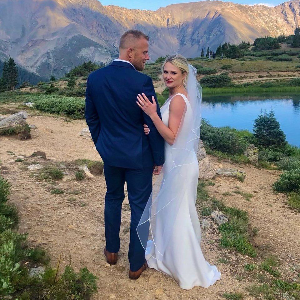 Wedding couple posing by a lake in the mountains. Bride in white dress, groom in blue suit, scenic background.