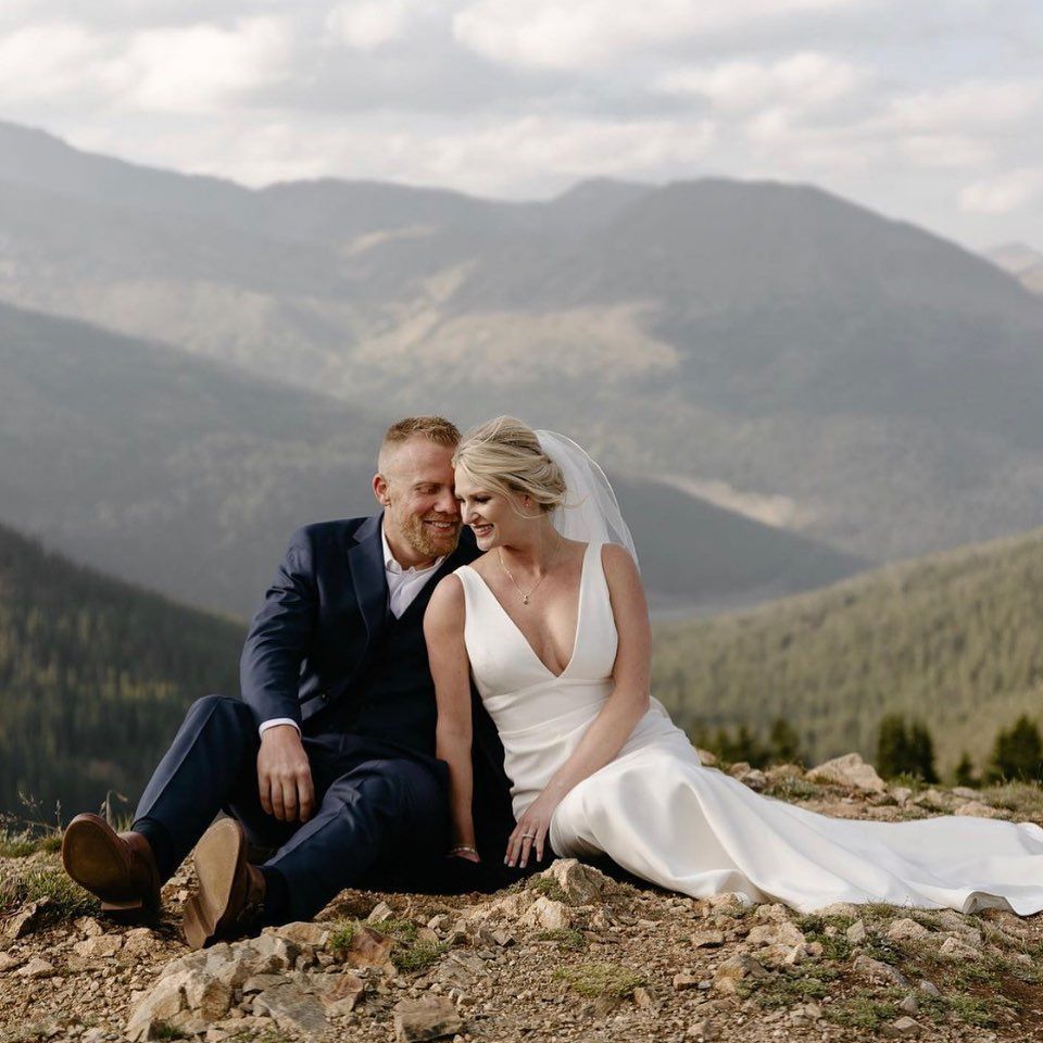 Bride and groom sit on a mountaintop, looking at each other. They're dressed in wedding attire