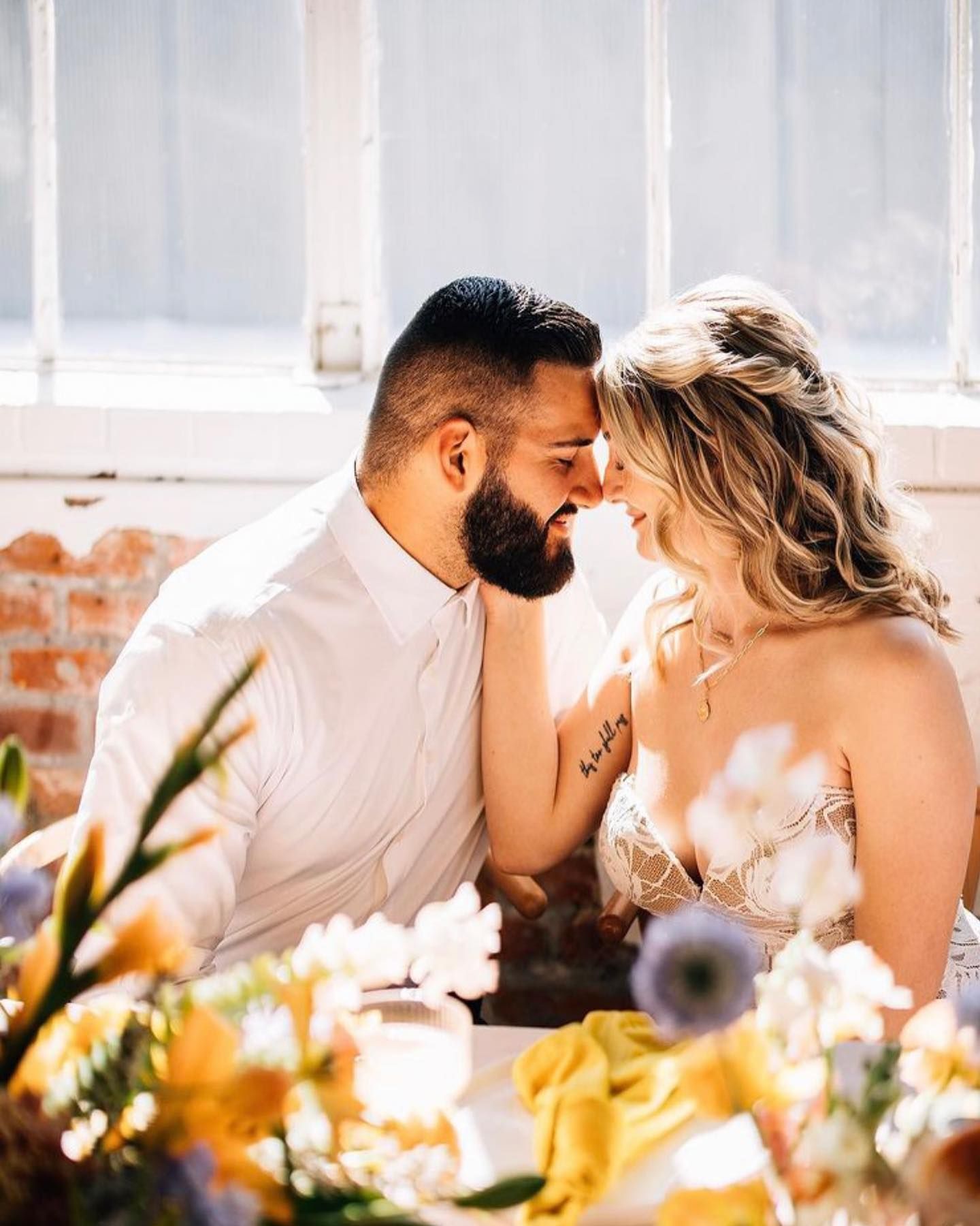 Couple touching foreheads, soft smiles, seated at a table with flowers. Sunlight streams through a window.
