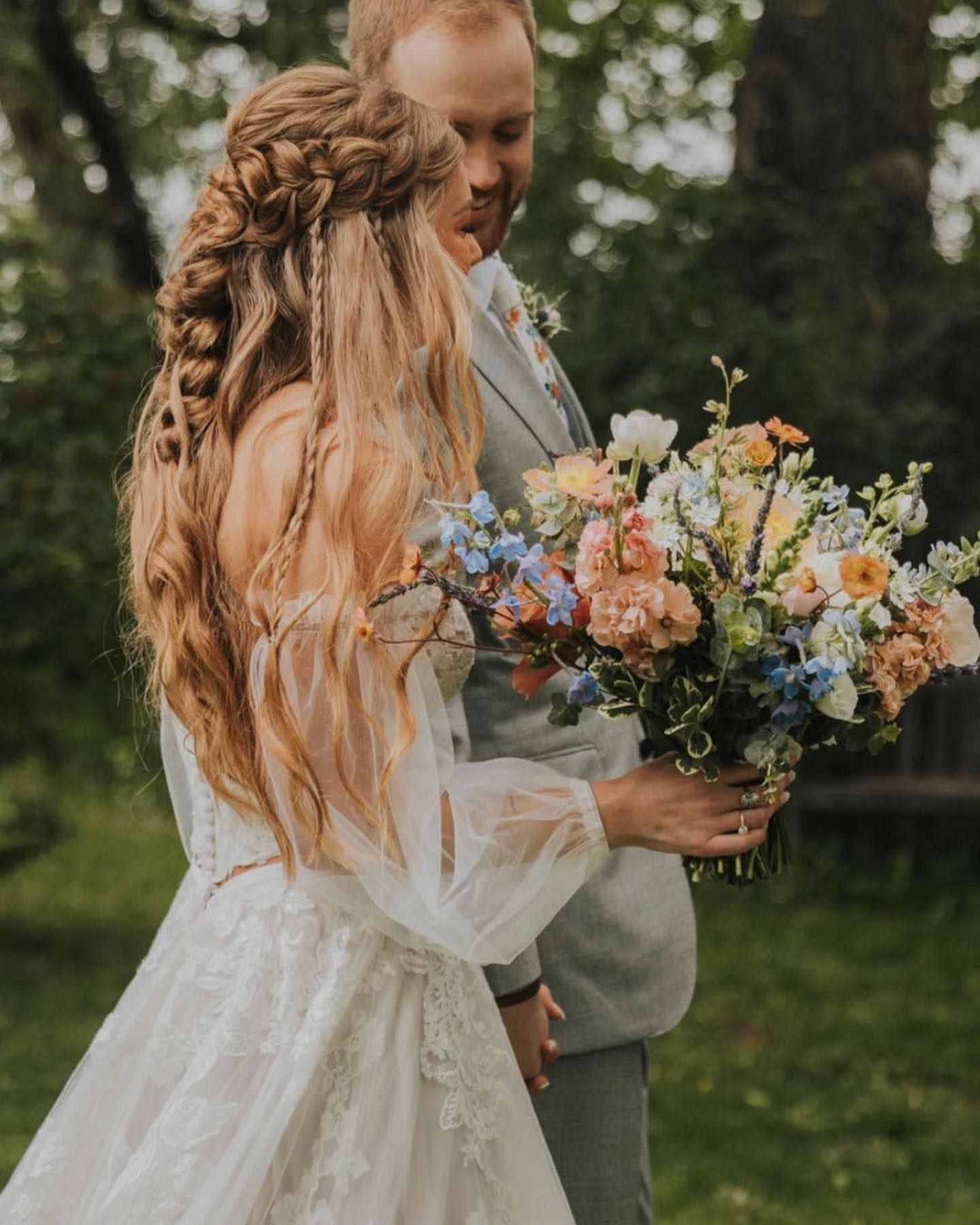 Bride in flowing white dress with floral bouquet, walking with groom in gray suit in a garden.
