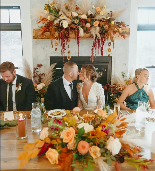 Wedding reception: couple kissing, seated at a table with colorful floral centerpiece, fireplace backdrop, guests nearby.