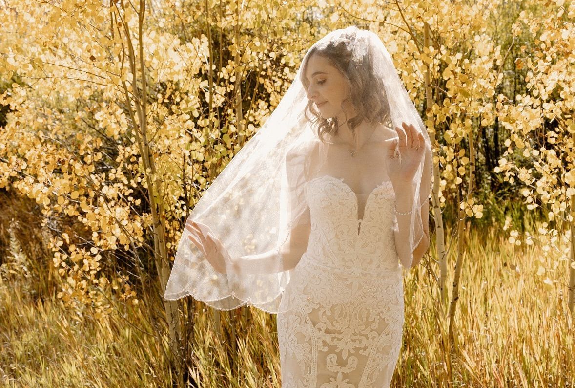 Bride in a lace wedding dress and veil outdoors, arms slightly outstretched, with fall foliage in the background.
