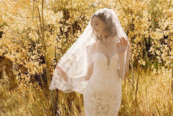 Bride in a lace wedding dress and veil outdoors, arms slightly outstretched, with fall foliage in the background.