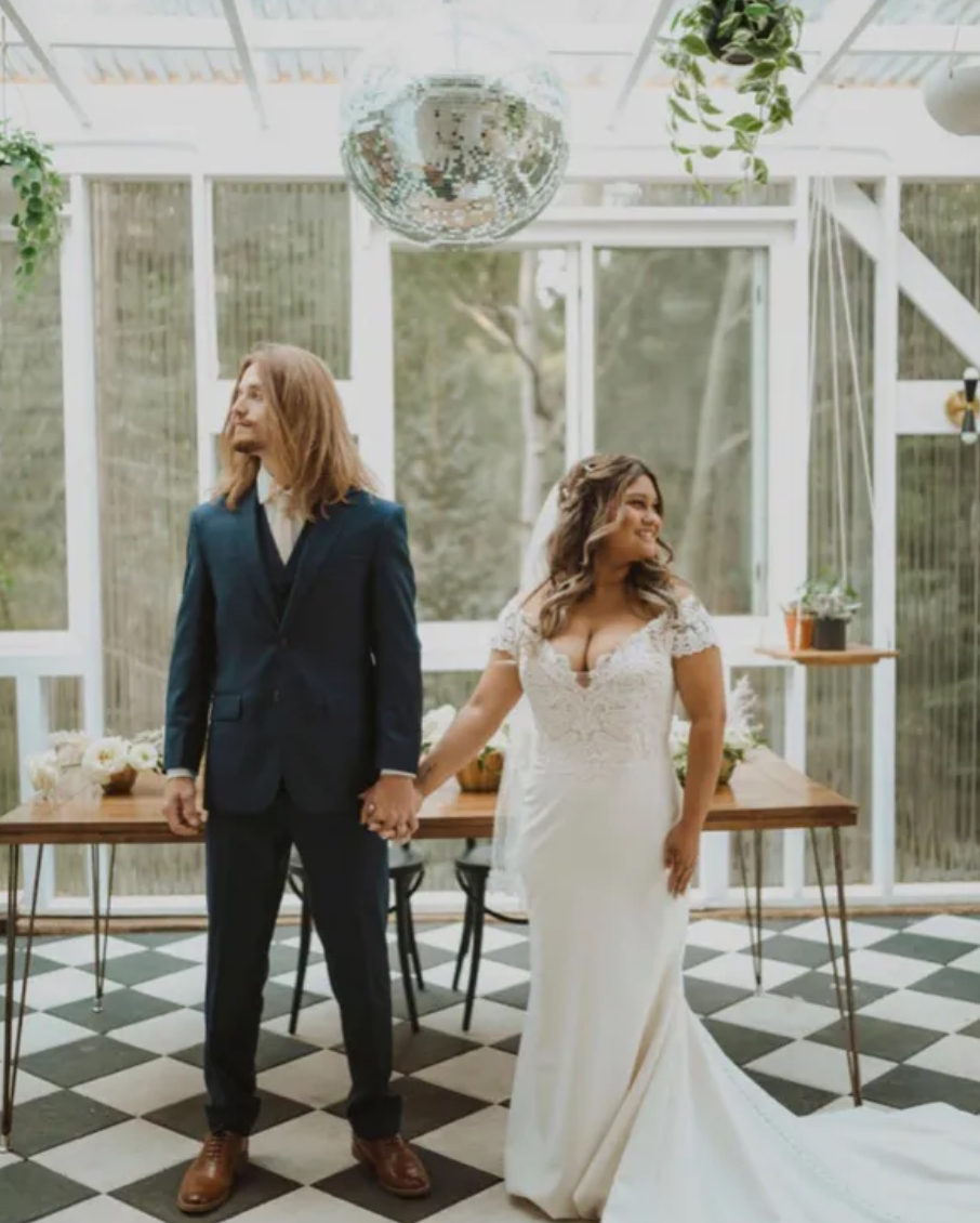 Newlyweds embrace on a rocky mountain, bride holding bouquet, groom kissing her forehead. Overcast sky.
