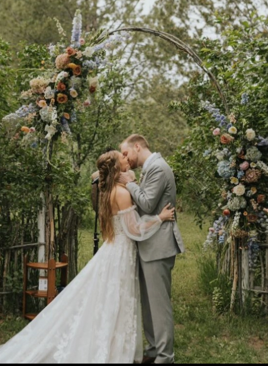 Bride in white gown looks at man in suit, holding bouquet, in a forest.