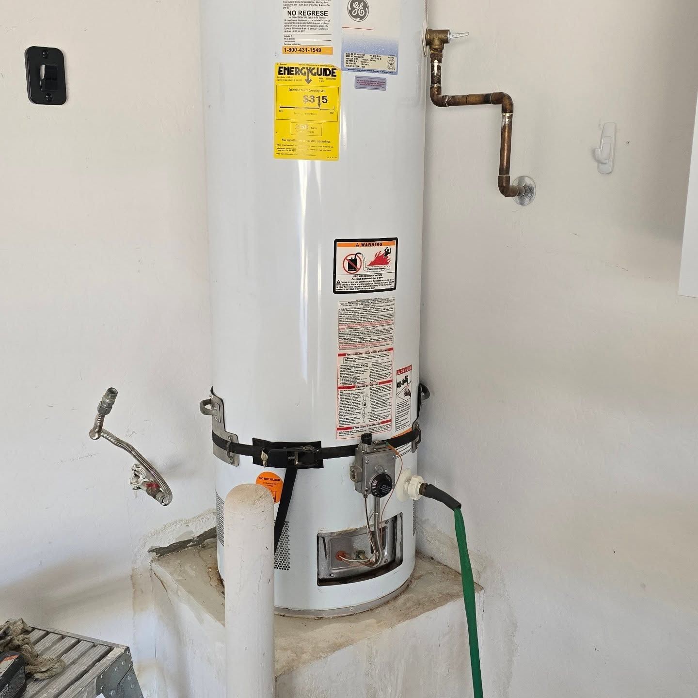 A white water heater is sitting on a shelf in a room next to a hose.