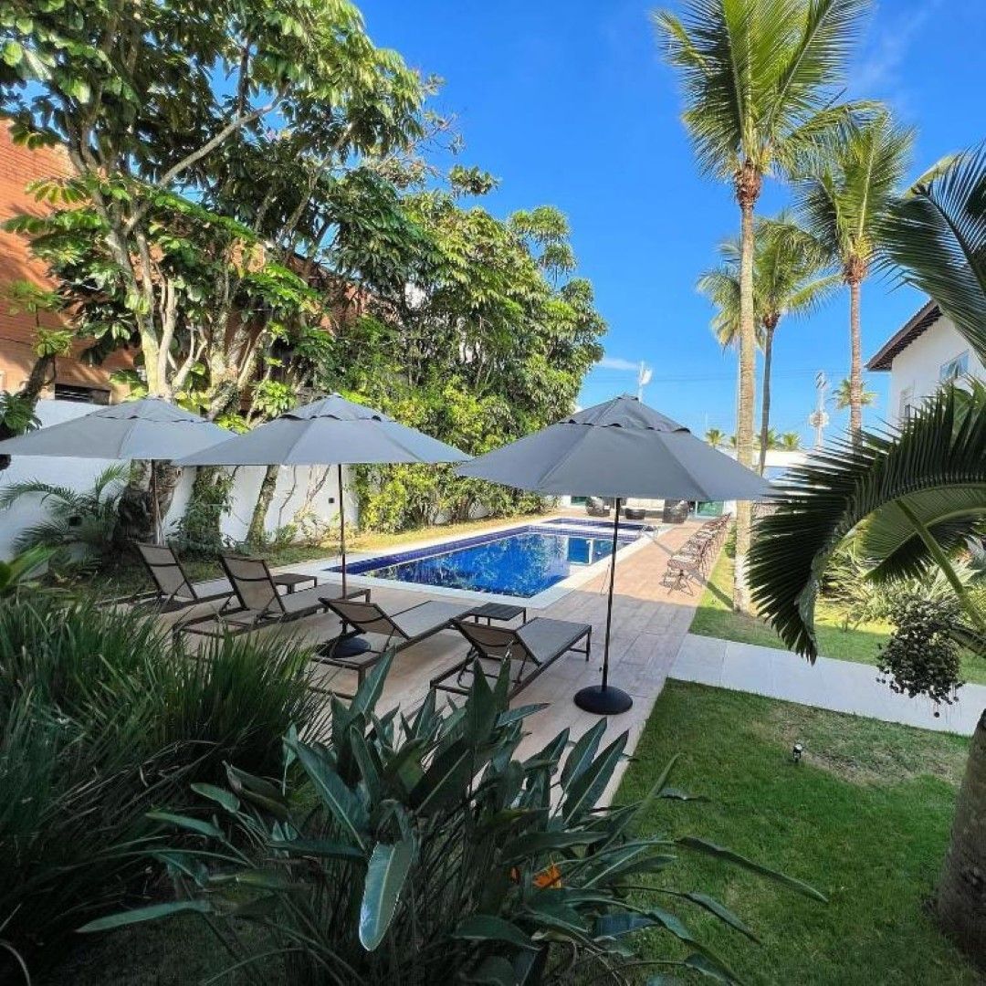 Piscina de resort tropical com guarda-sóis cinzentos, espreguiçadeiras, palmeiras e céu azul.