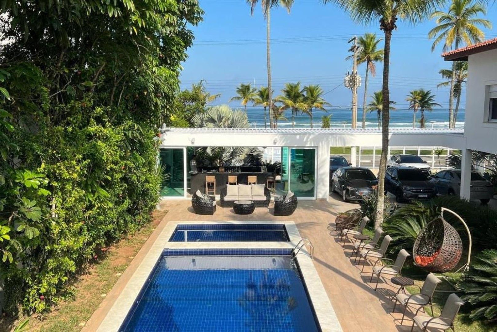 Piscina de resort tropical com espreguiçadeiras, palmeiras e vista para o mar além do pátio.