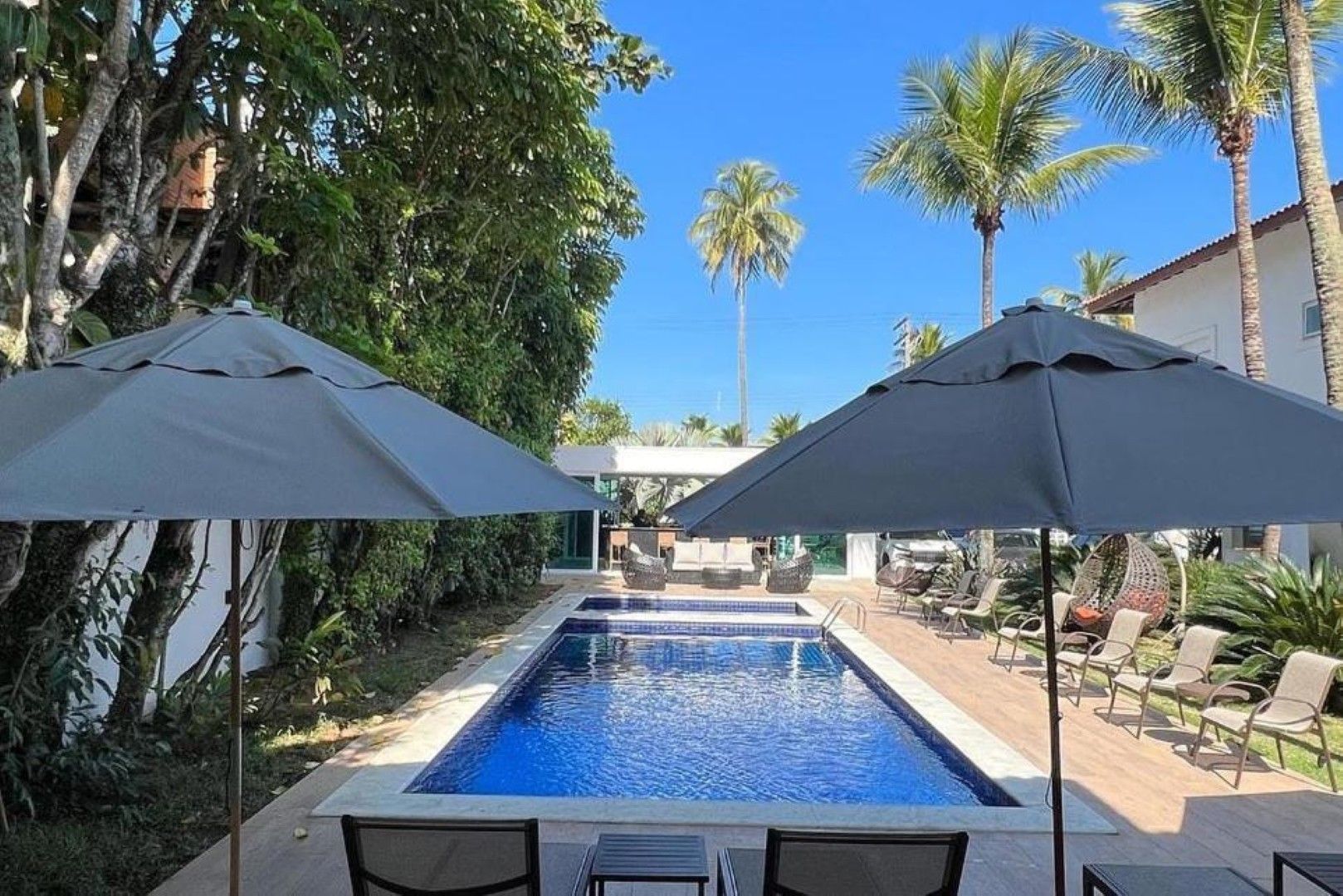 Pátio à beira da piscina com piscina azul, guarda-sóis pretos e espreguiçadeiras em um pátio tropical.