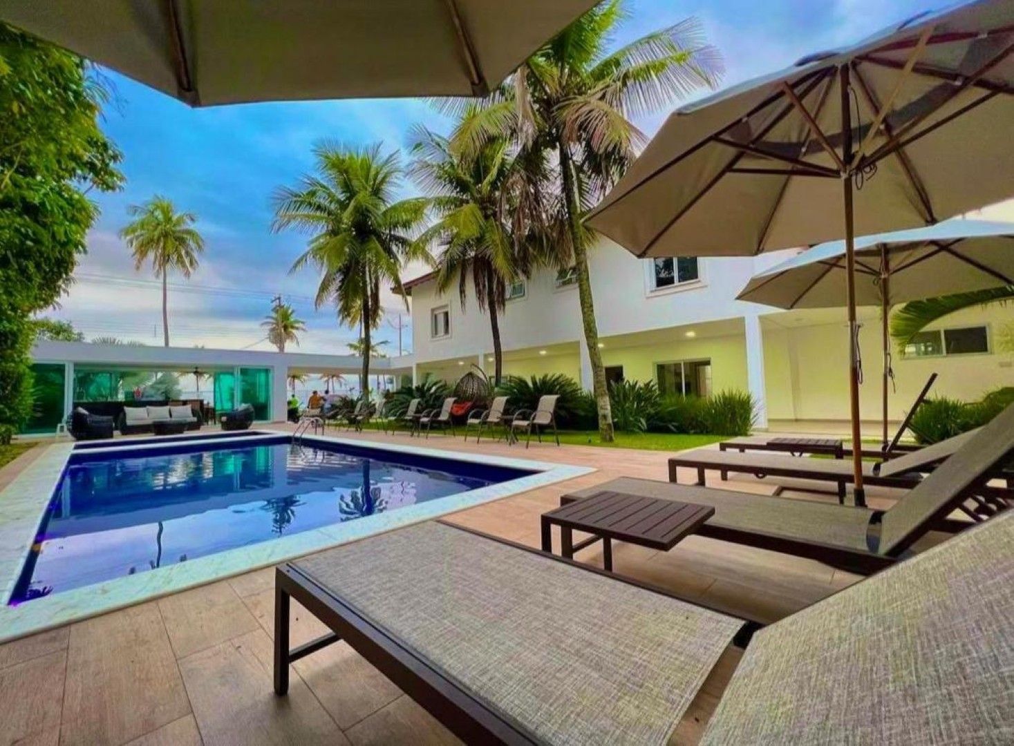 Piscina do resort com espreguiçadeiras, guarda-sóis, palmeiras e vista para o mar sob um céu azul brilhante.