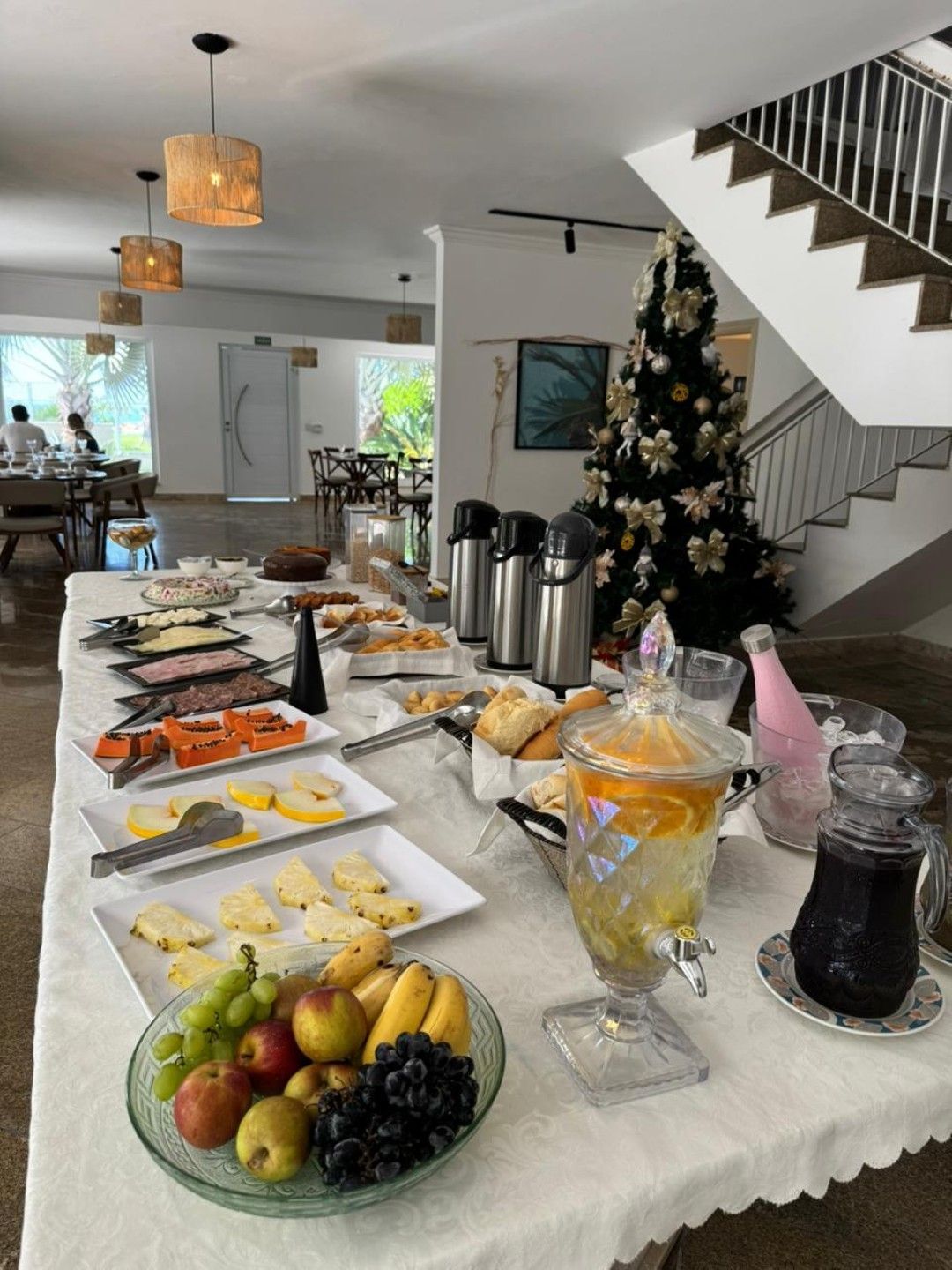 Mesa de buffet com pratos de café da manhã e frutas, ao lado de uma árvore de Natal decorada em uma casa iluminada.