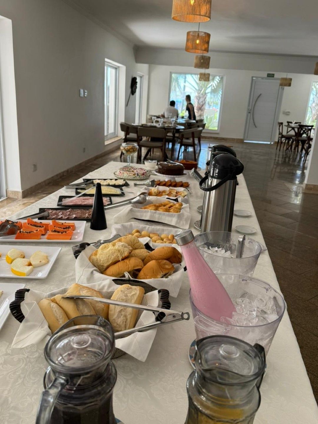 Mesa de buffet com doces, frutas, café e jarras em uma sala de jantar iluminada.