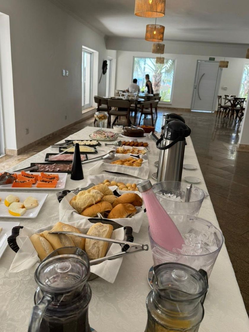 Mesa de buffet com doces, frutas, café e jarras em uma sala de jantar iluminada.