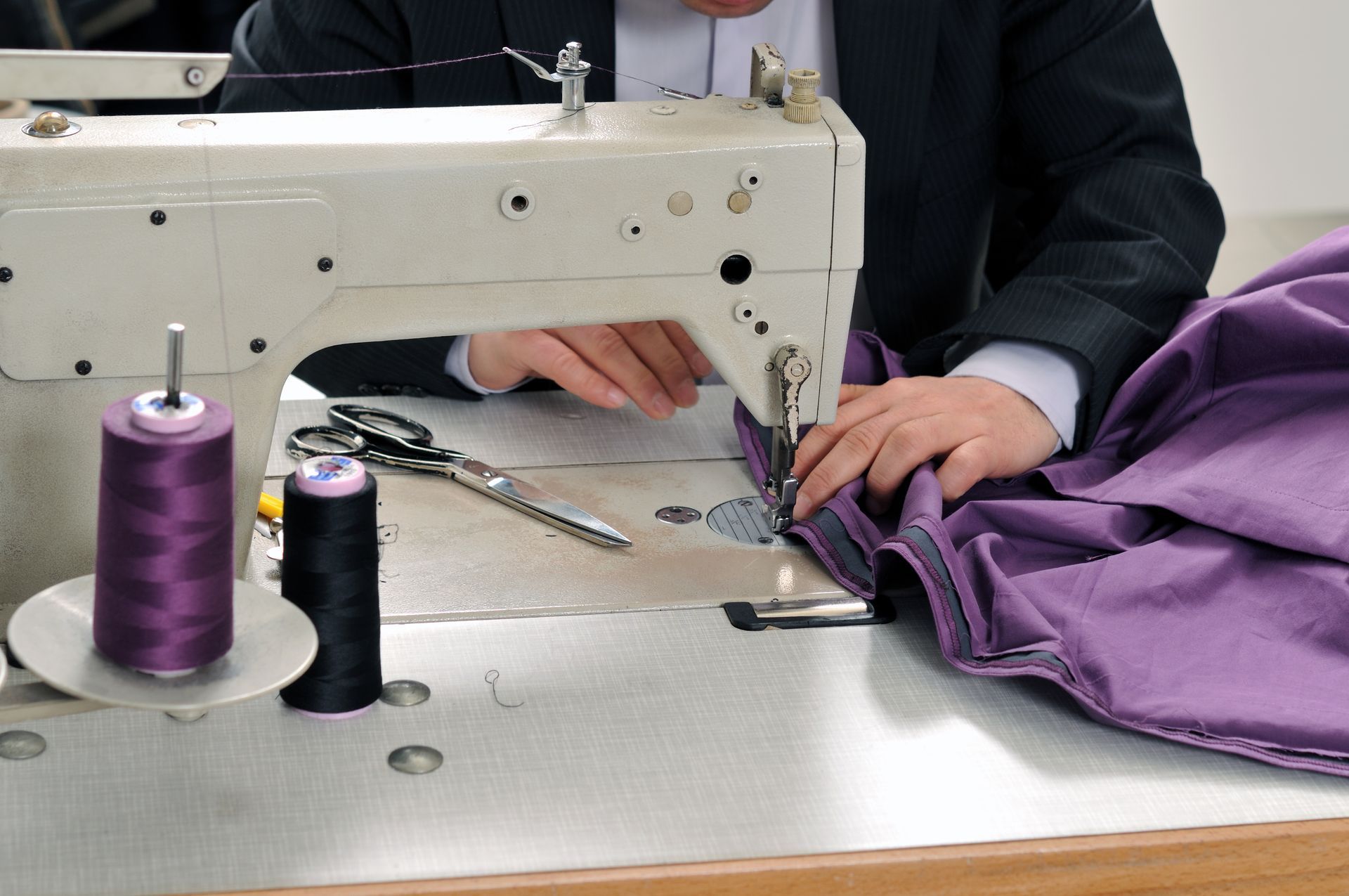 A man in a suit is using a sewing machine to sew a piece of purple fabric