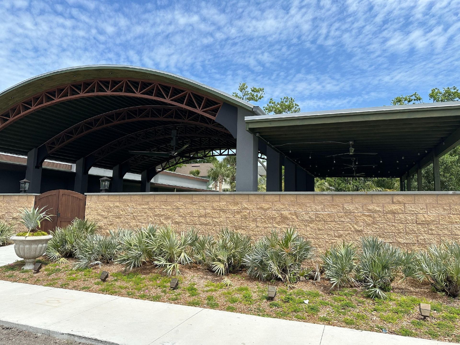 Outdoor event venue with a large arched roof, open-air stage, and brick wall with greenery. Blue sky in the background.