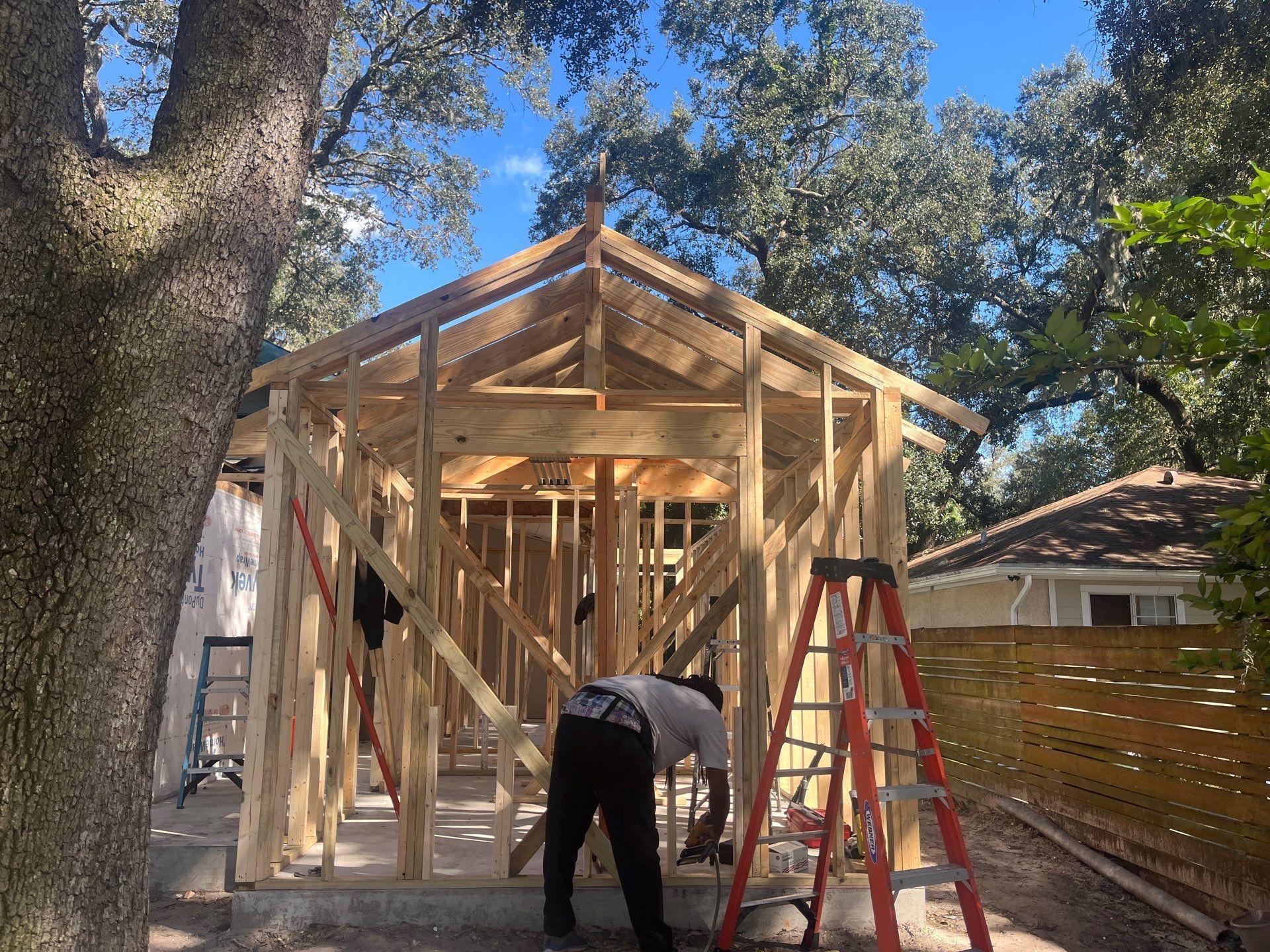A person building a wooden structure outdoors with a ladder and surrounding trees. Bright blue sky overhead.