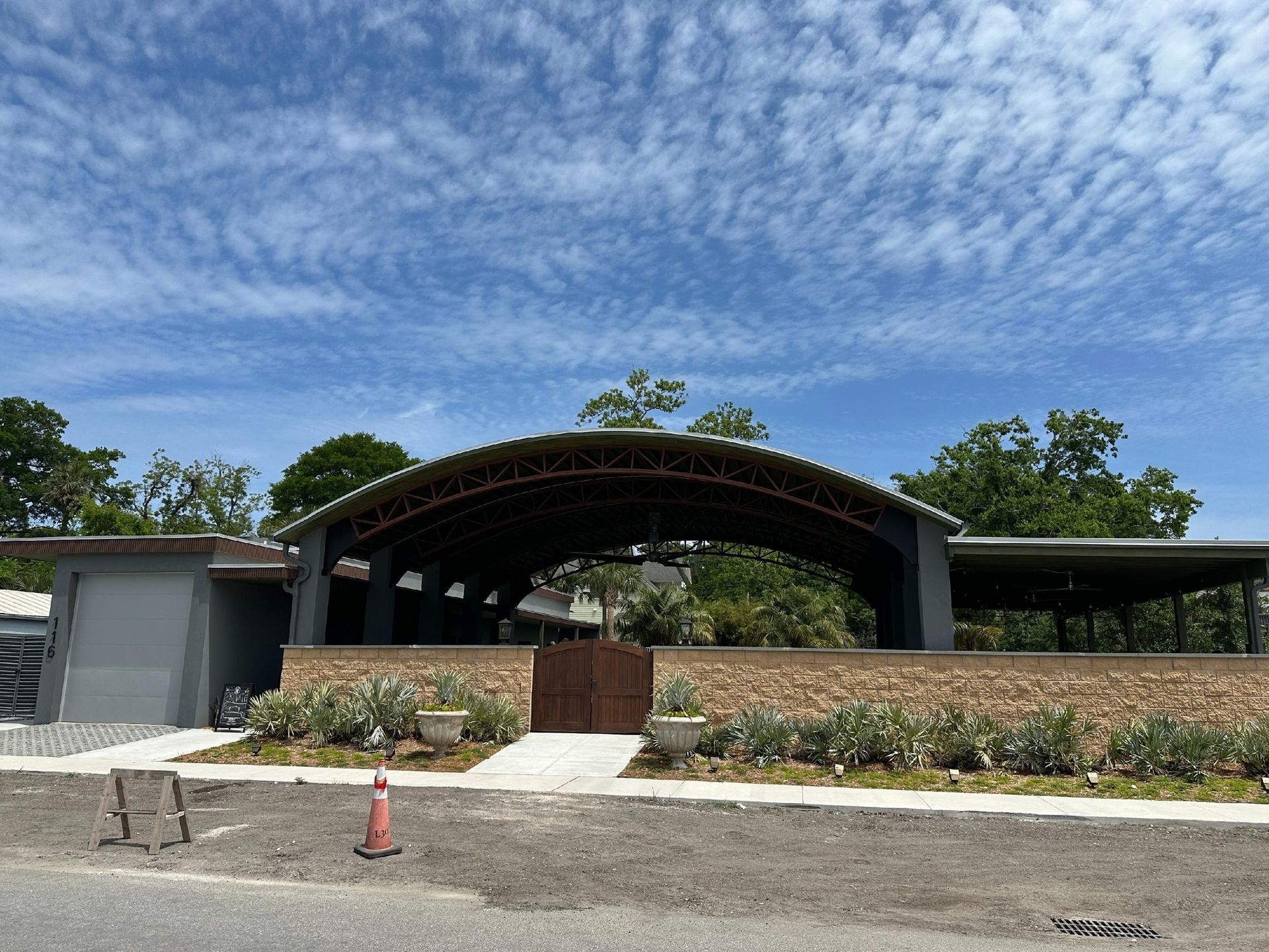 Building with arched roof, tan wall, and wooden gate under a cloudy blue sky. Trees and shrubs surround.