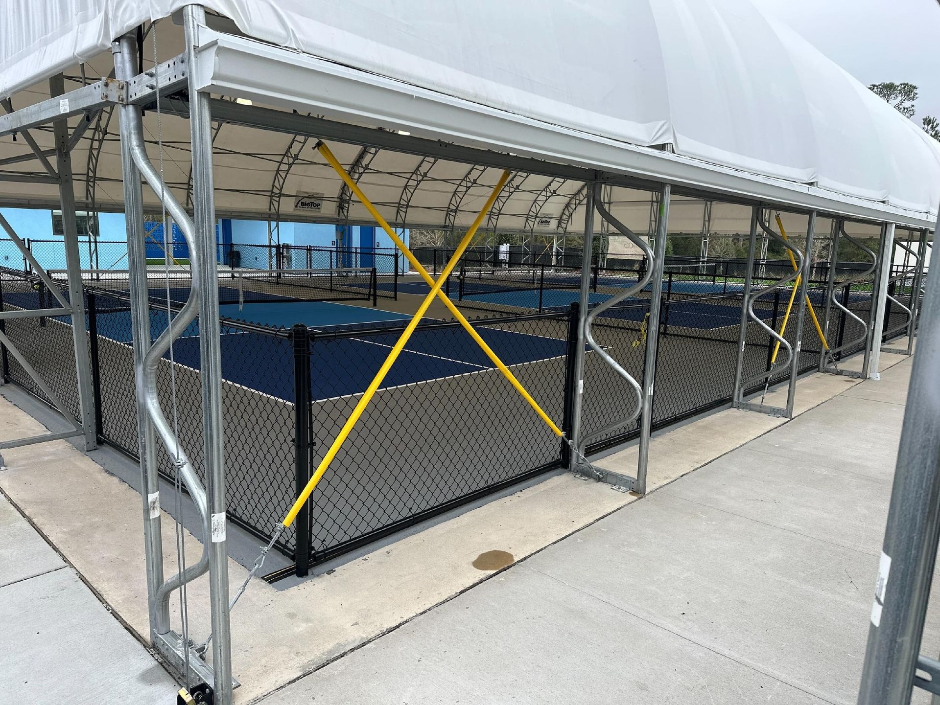A covered pickleball court with a white tent roof, surrounded by a black fence and supported by metal beams.
