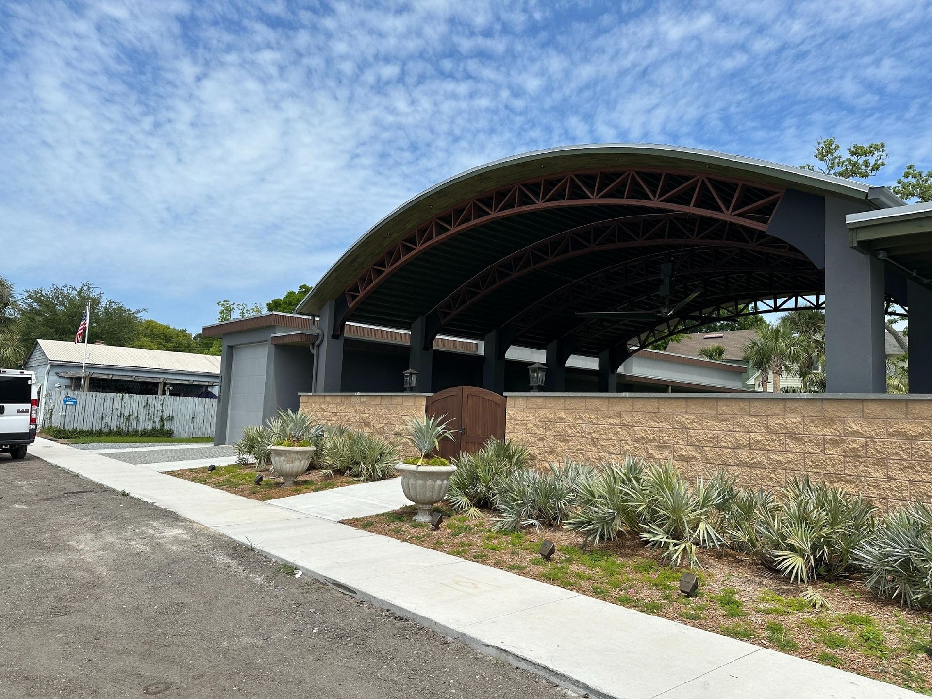 Outdoor pavilion with curved roof, surrounded by landscaping and concrete. Features include gray structure, brown and green plants, and blue sky.