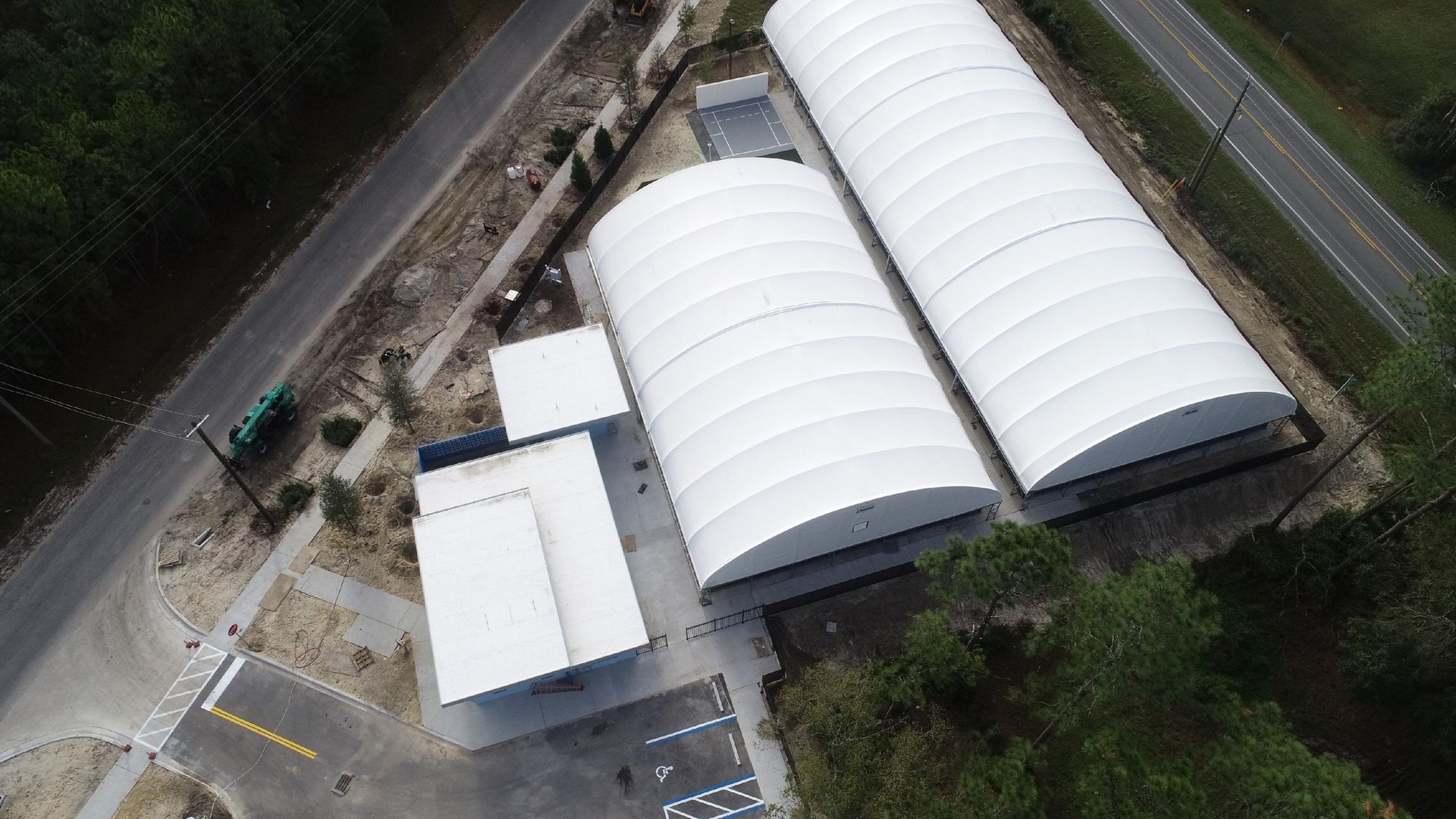 Overhead view of a white arched-roof building complex next to a road and trees. The buildings are surrounded by concrete and asphalt.