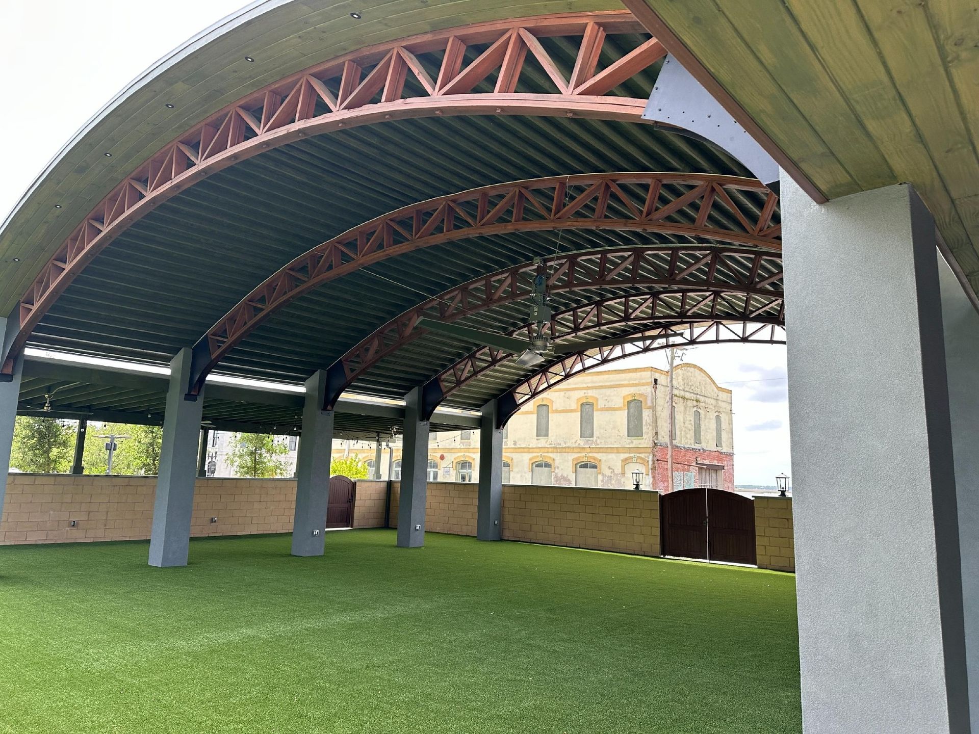 Covered outdoor area with artificial green turf floor, brown arched beams, gray columns, and a building visible in the background.