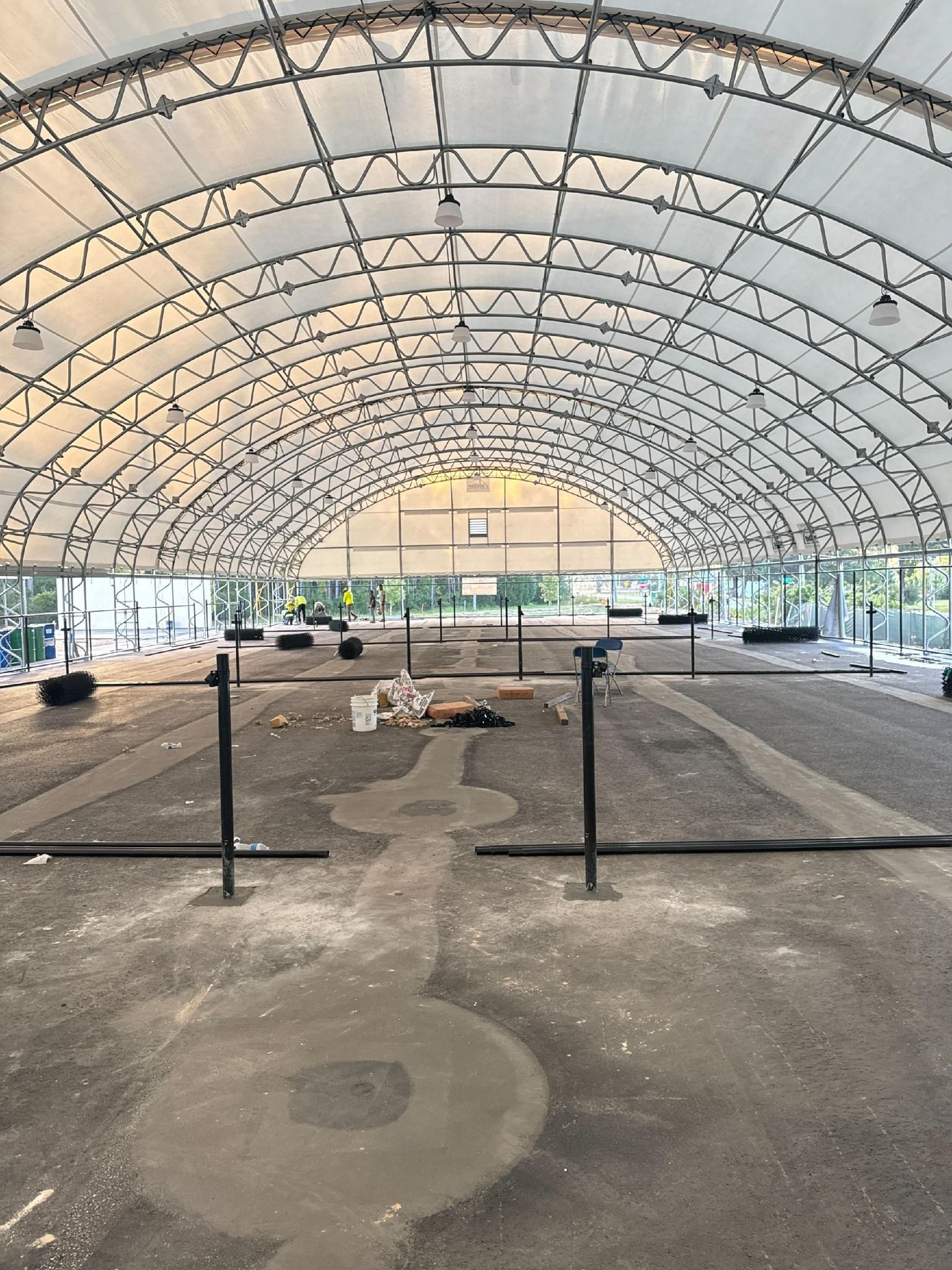 Interior of a large, white-roofed structure, possibly a sports hall. Construction is in progress; gray floor with areas of concrete.