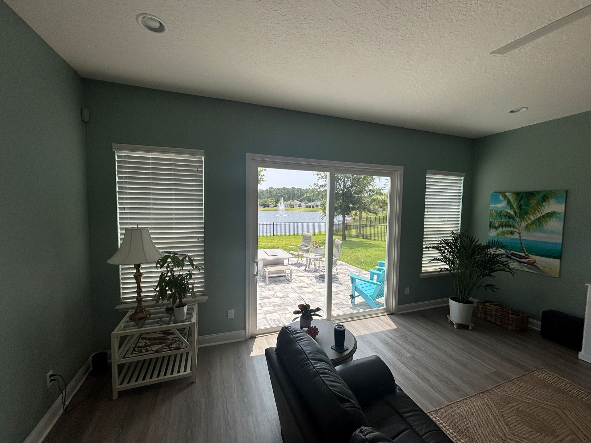 A living room with teal walls and a large sliding glass door overlooking a patio and lake.  Furniture includes a black couch and wicker table.
