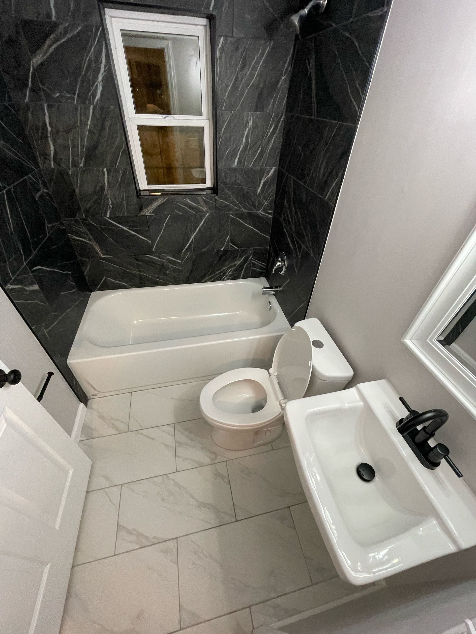 A newly renovated bathroom with white fixtures, gray and black marble-look wall tile, and gray floor tiles. A window is above the bathtub.