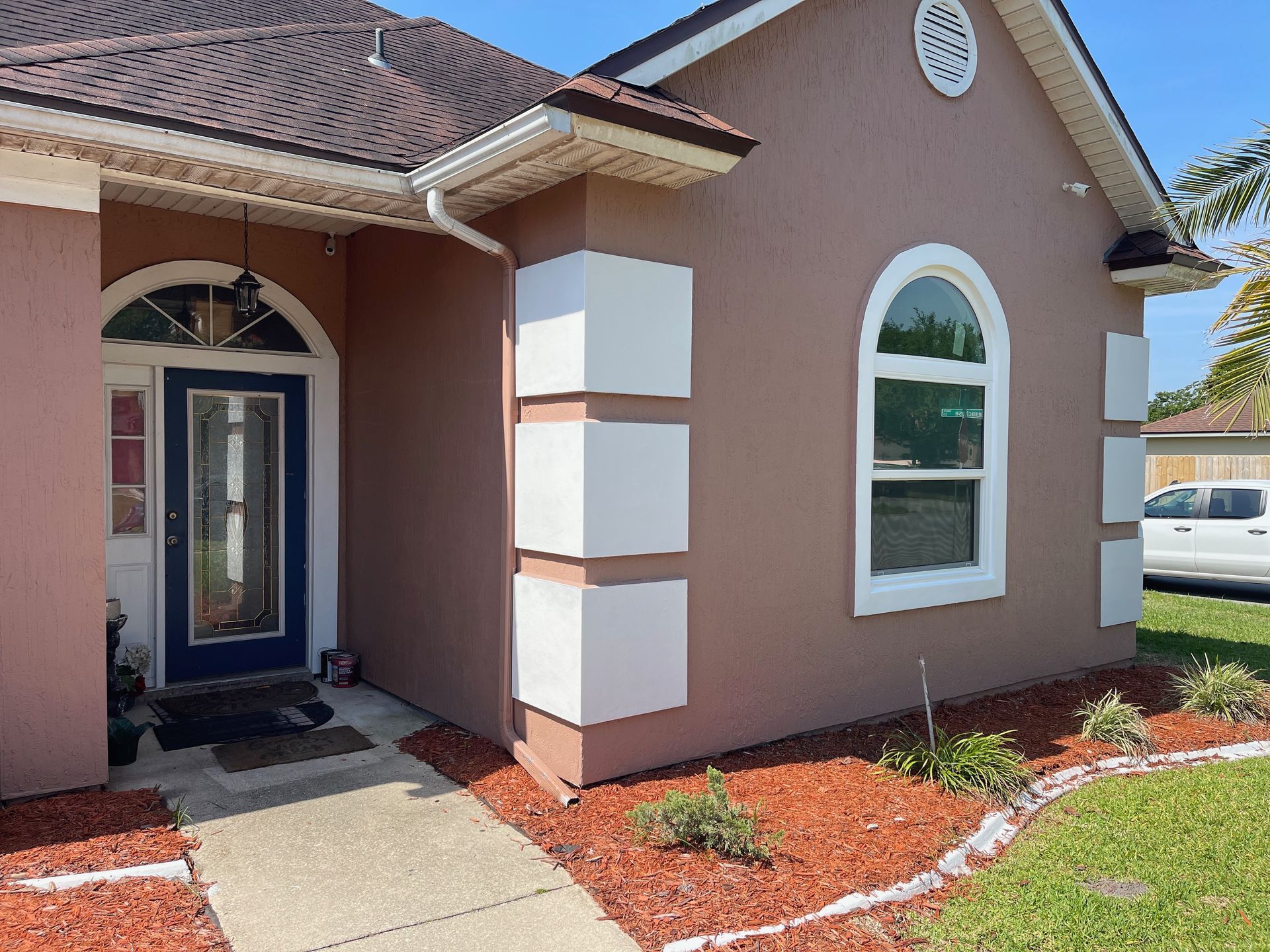 A pink stucco house with white trim. Features include a blue front door, arched window, and a walkway with red mulch.