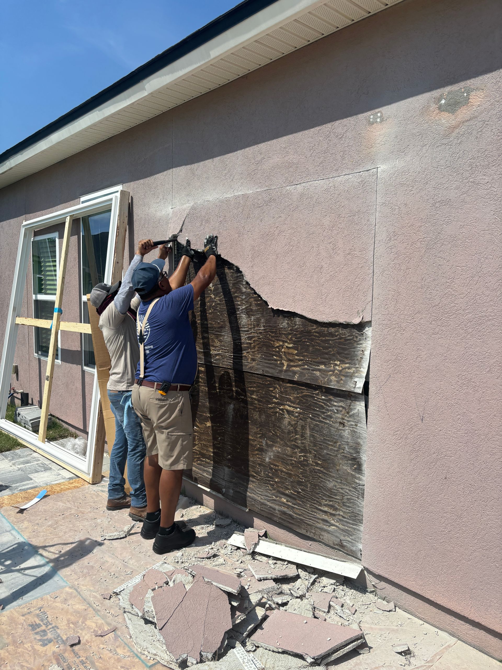 Three construction workers removing a section of exterior wall from a building on a sunny day.